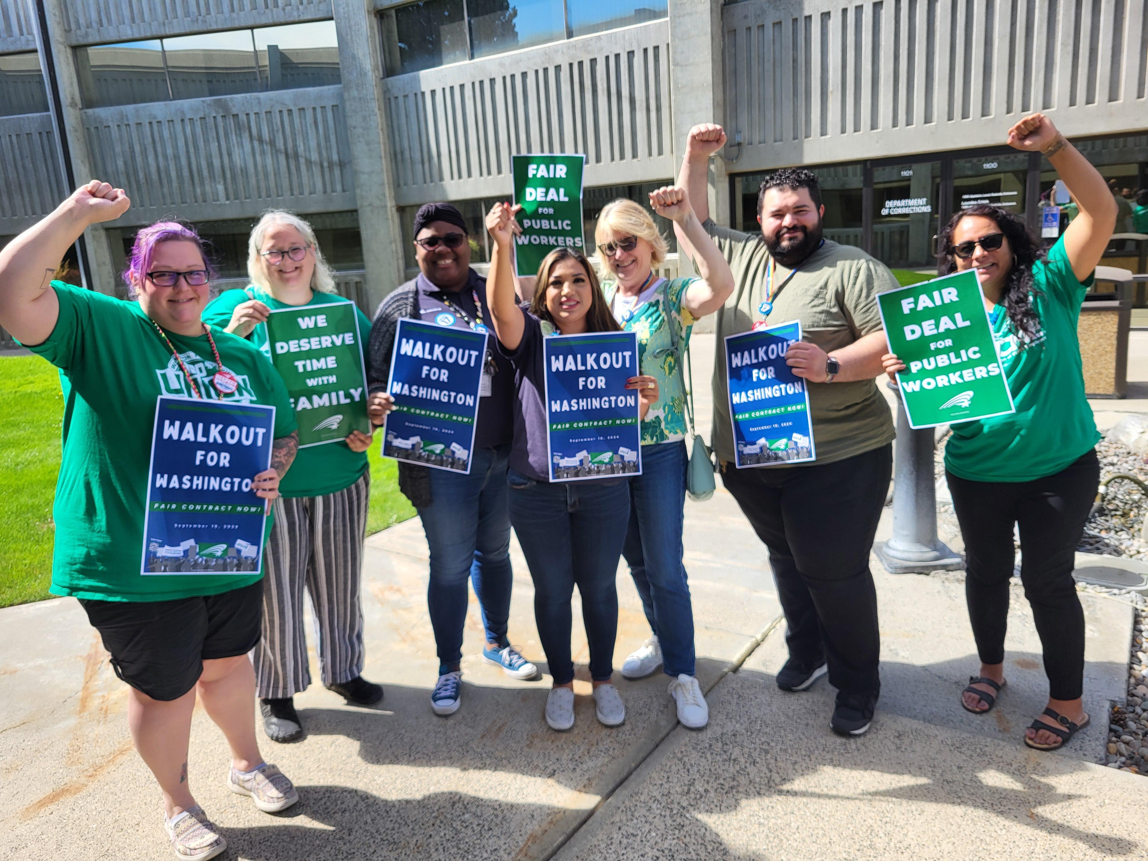 A group of seven WFSE DOC members stand outside in the daylight, some with fists raised. They are holding signs with WFSE branding that say "Public Workers United for Washington."